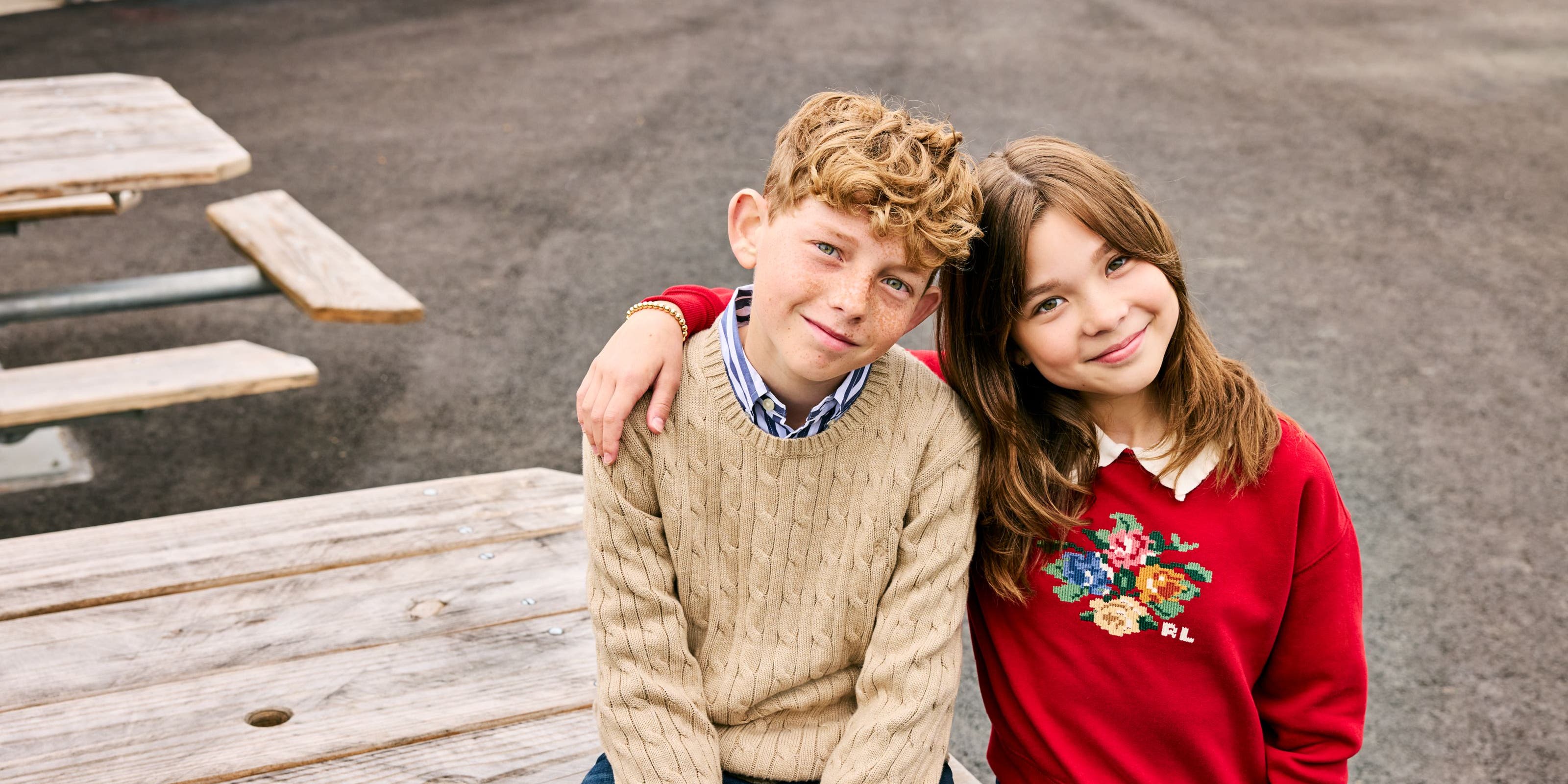 Two kids, a boy and a girl, sit together on a picnic table wearing cozy knit and cross-stitch sweaters.