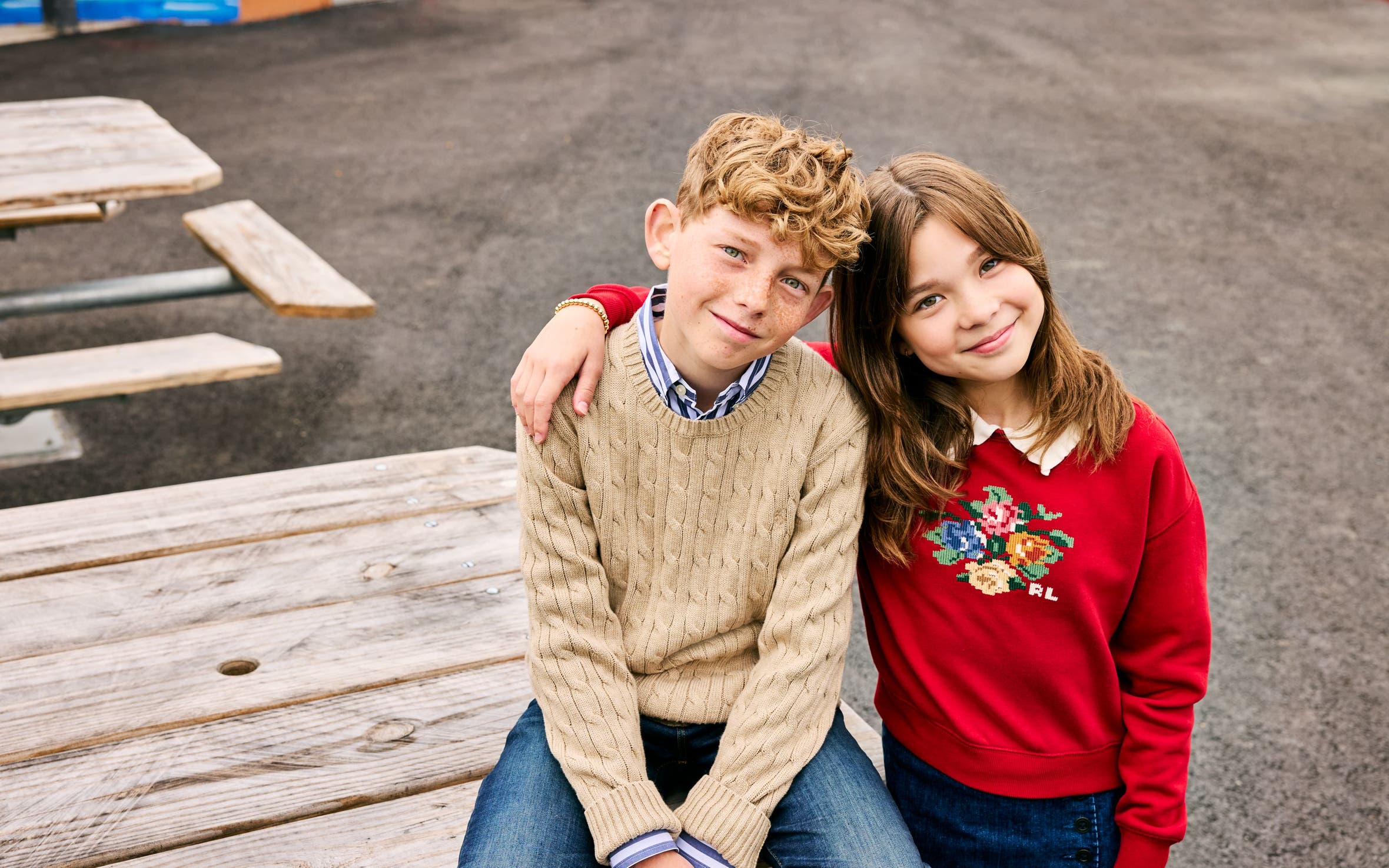 Two kids, a boy and a girl, sit together on a picnic table wearing cozy knit and cross-stitch sweaters.