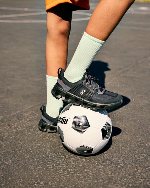 Close-up of a kid's legs in mint green socks and black On sneakers, with one foot resting on a soccer ball on a paved court.