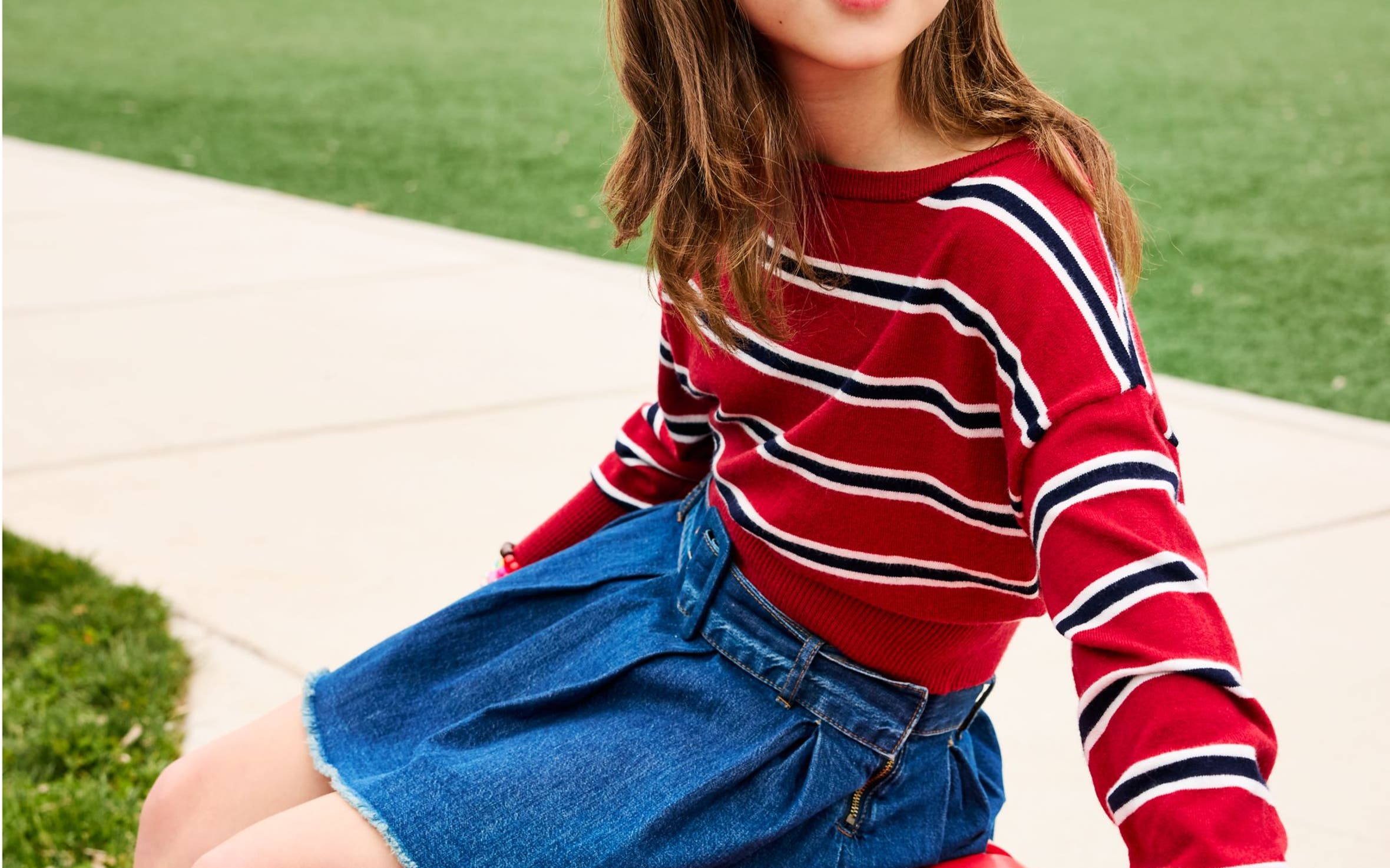 Girl in a red, navy and white striped long-sleeve top and denim skirt sitting on a red bench.