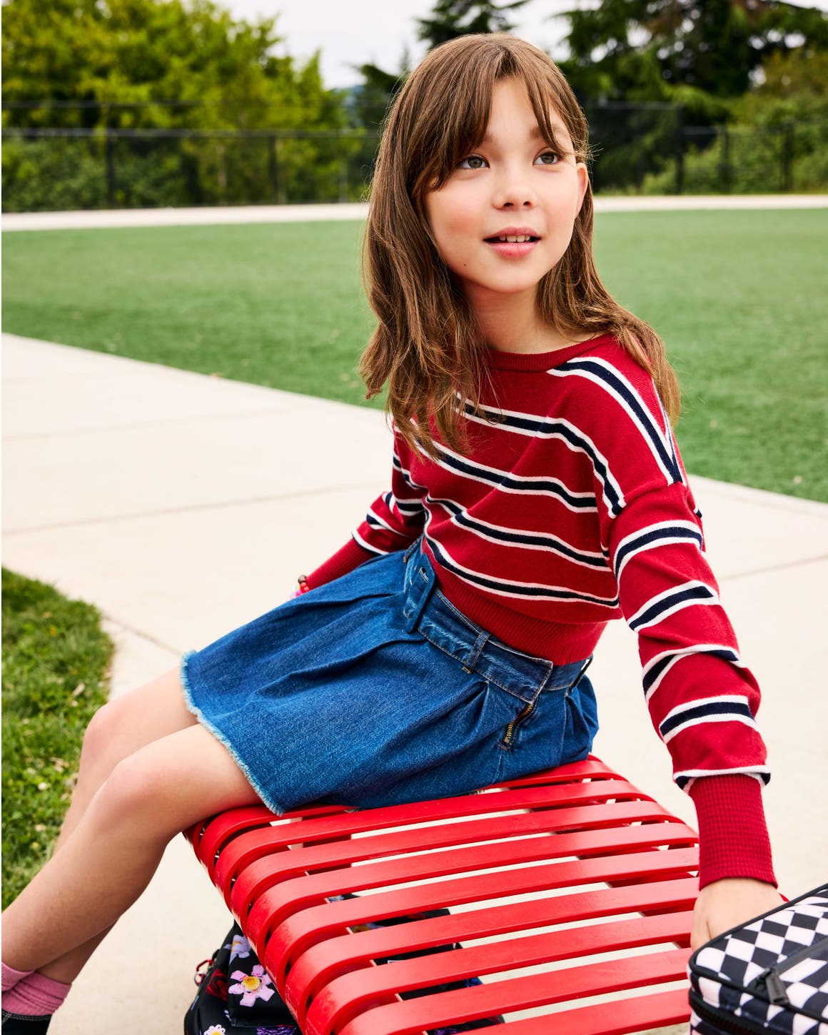 Girl in a red, navy and white striped long-sleeve top and denim skirt sitting on a red bench.