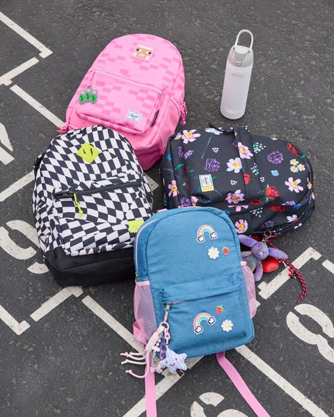 Four backpacks — pink with patches, black and white checkered, floral print and blue with a rainbow patch — arranged on pavement with a water bottle.