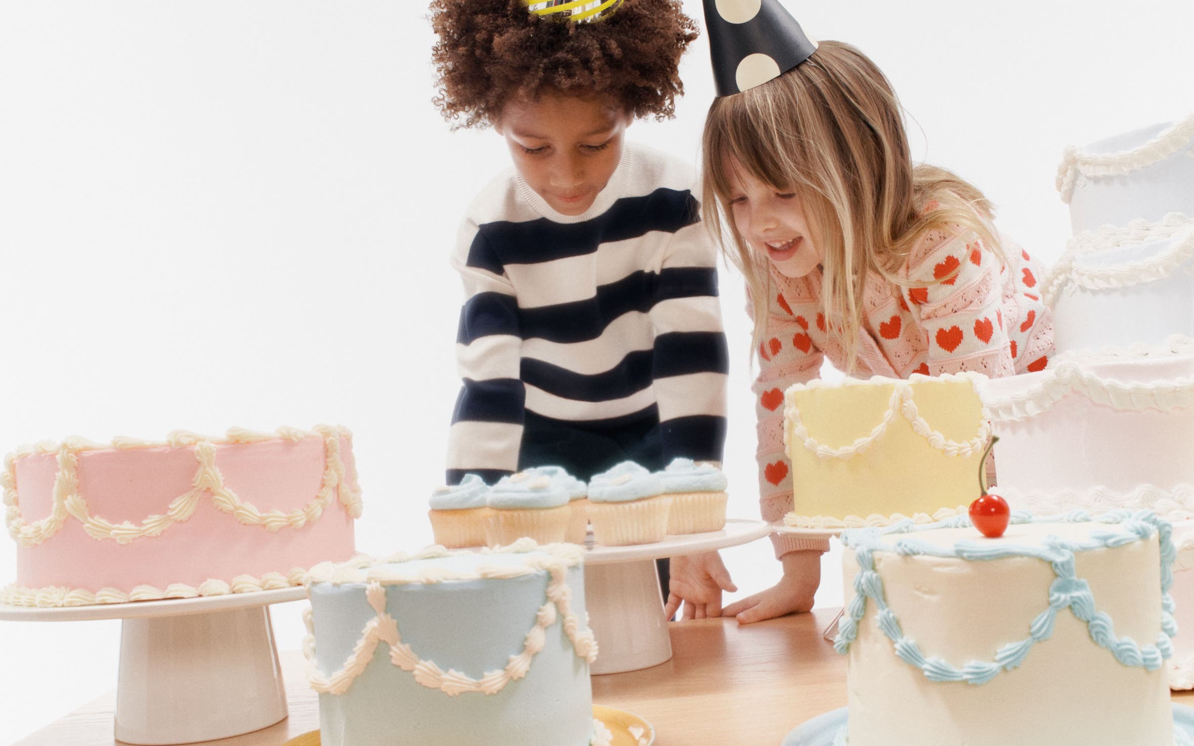 A boy and girl in party hats looking at birthday cakes.