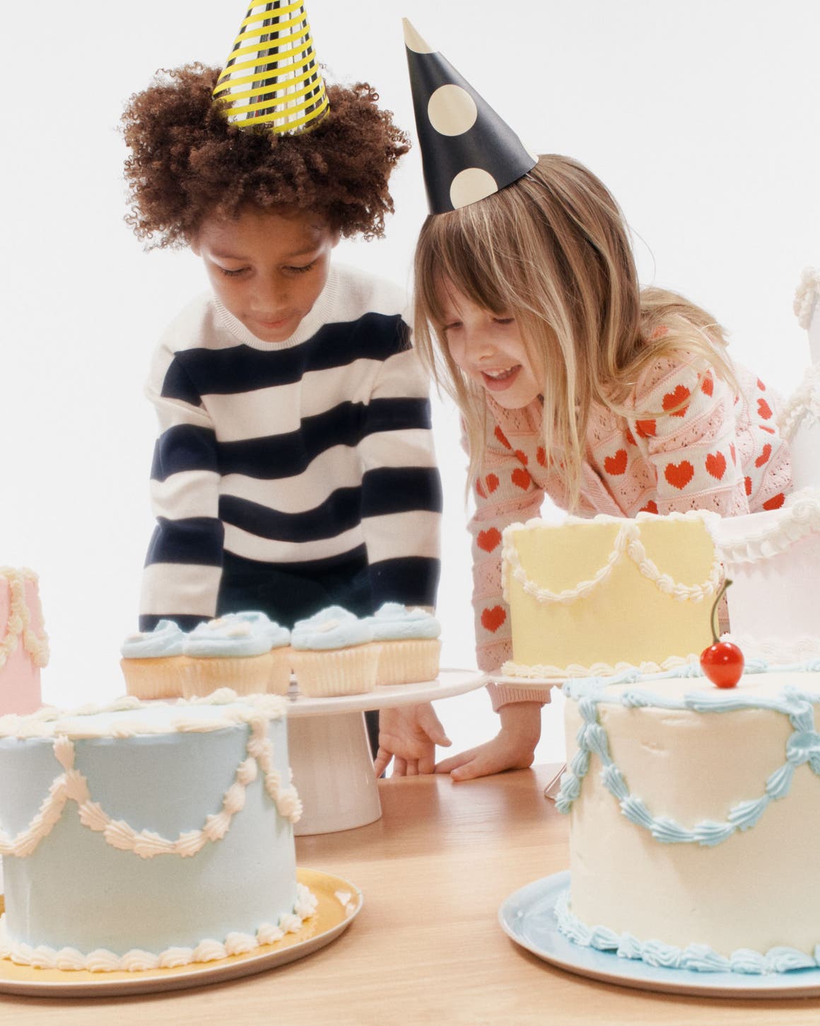 A boy and girl in party hats looking at birthday cakes.