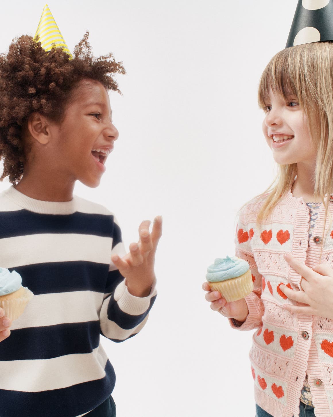 A boy and girl in sale styles wearing party hats and eating cupcakes.
