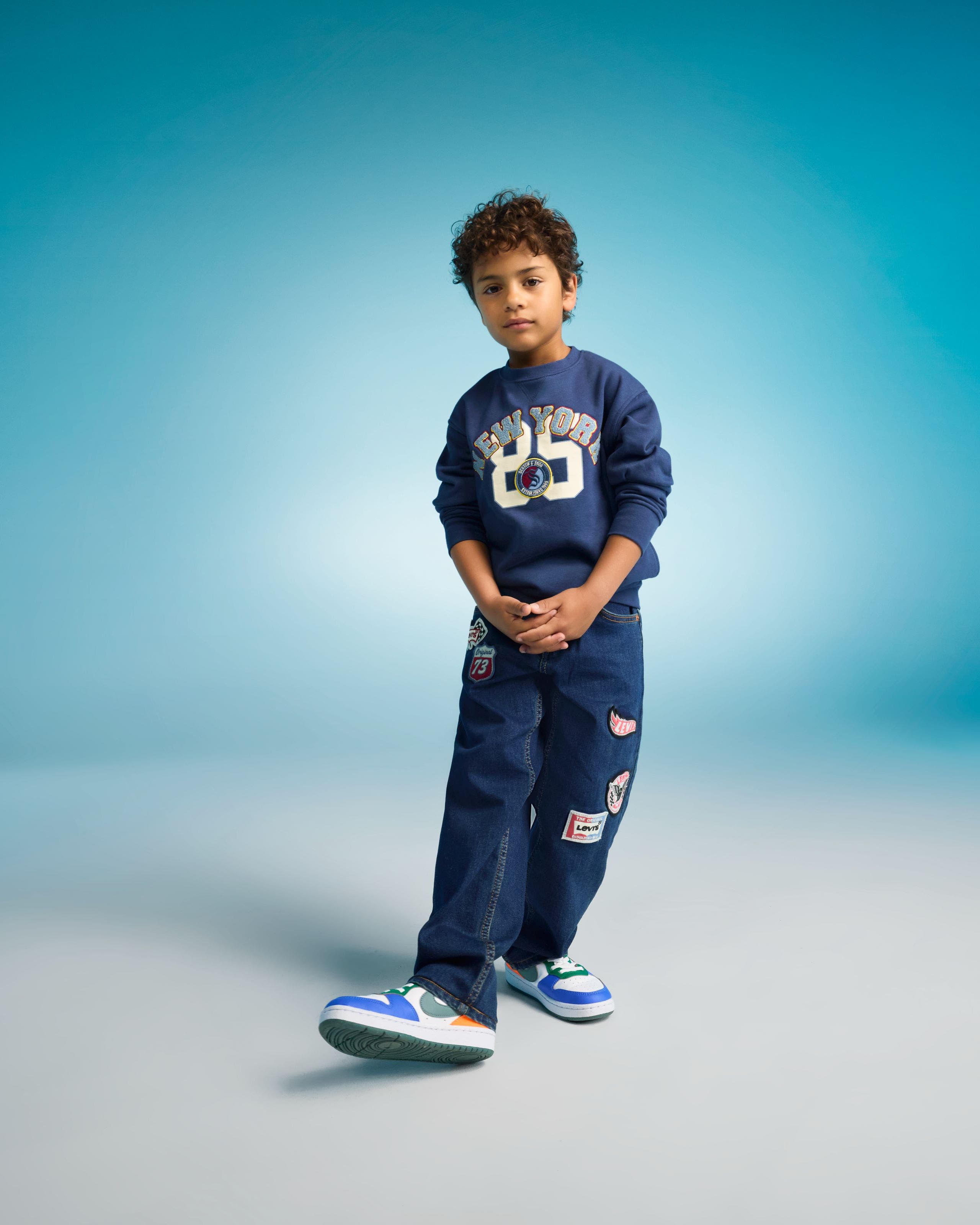 Boy wearing a navy sweatshirt with a “25” graphic, matching dark jeans with patches, and blue-and-white sneakers.
