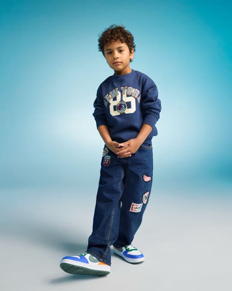 Boy wearing a navy sweatshirt with a “25” graphic, matching dark jeans with patches, and blue-and-white sneakers.