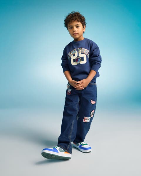 Boy model wearing a navy graphic sweatshirt and dark jeans with blue and white sneakers.