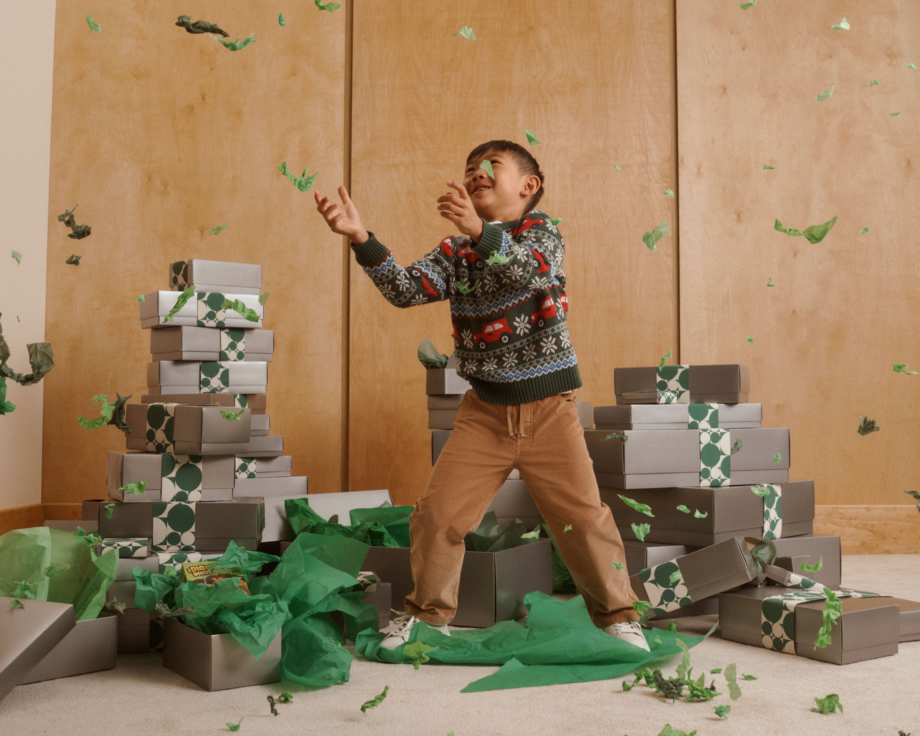 Boy in a holiday sweater tossing green tissue confetti among stacks of gift boxes.