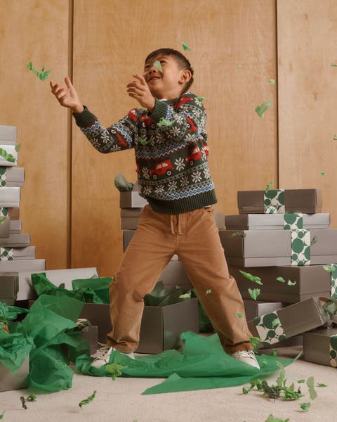 Boy in a holiday sweater tossing green tissue confetti among stacks of gift boxes.