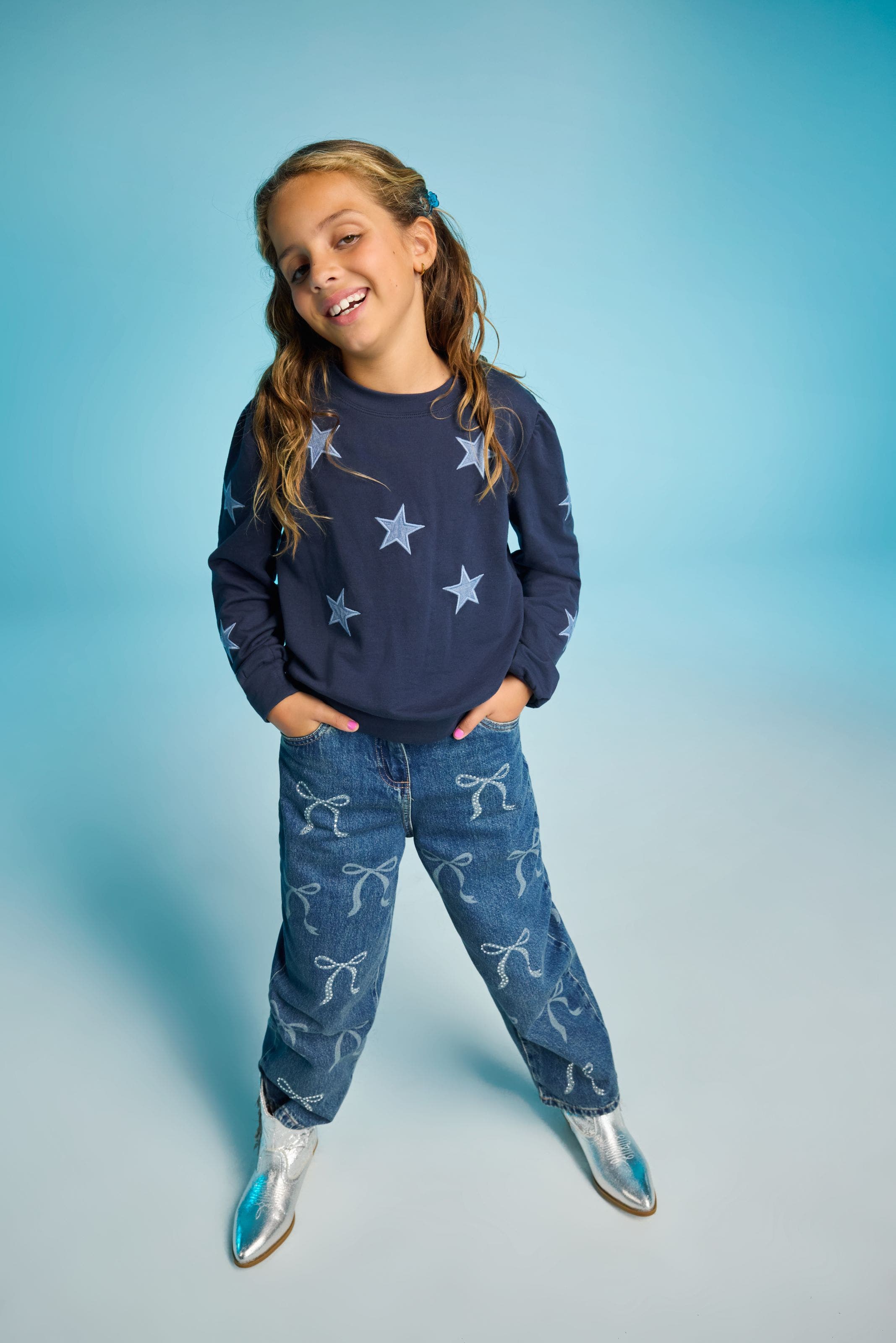 A child wearing a navy star print sweatshirt and bow printed jeans with silver shoes.