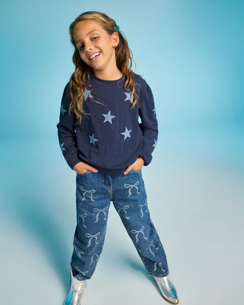 A child wearing a navy star print sweatshirt and bow printed jeans with silver shoes.