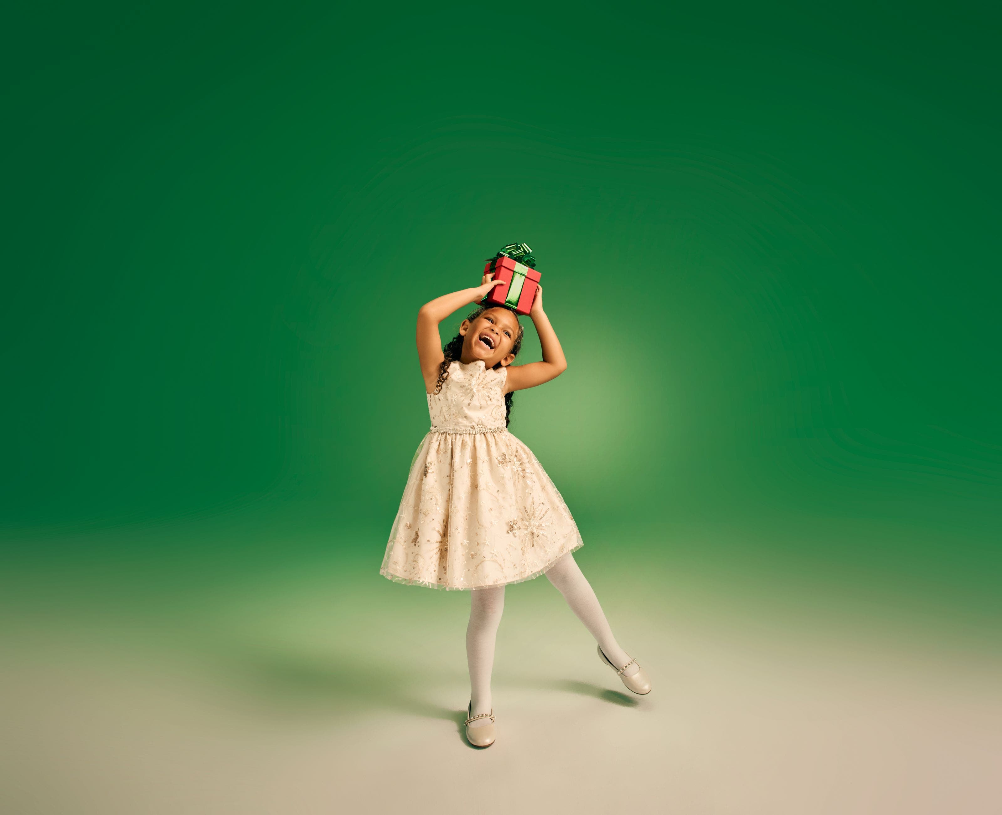 Young girl in a cream holiday dress balancing a red gift box on her head against a green background.