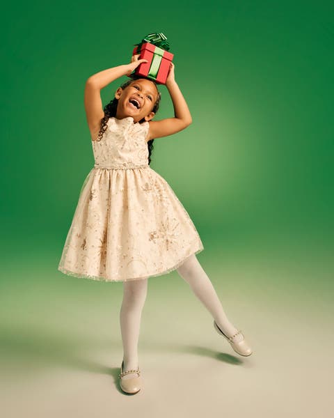 Young girl in a cream holiday dress balancing a red gift box on her head against a green background.