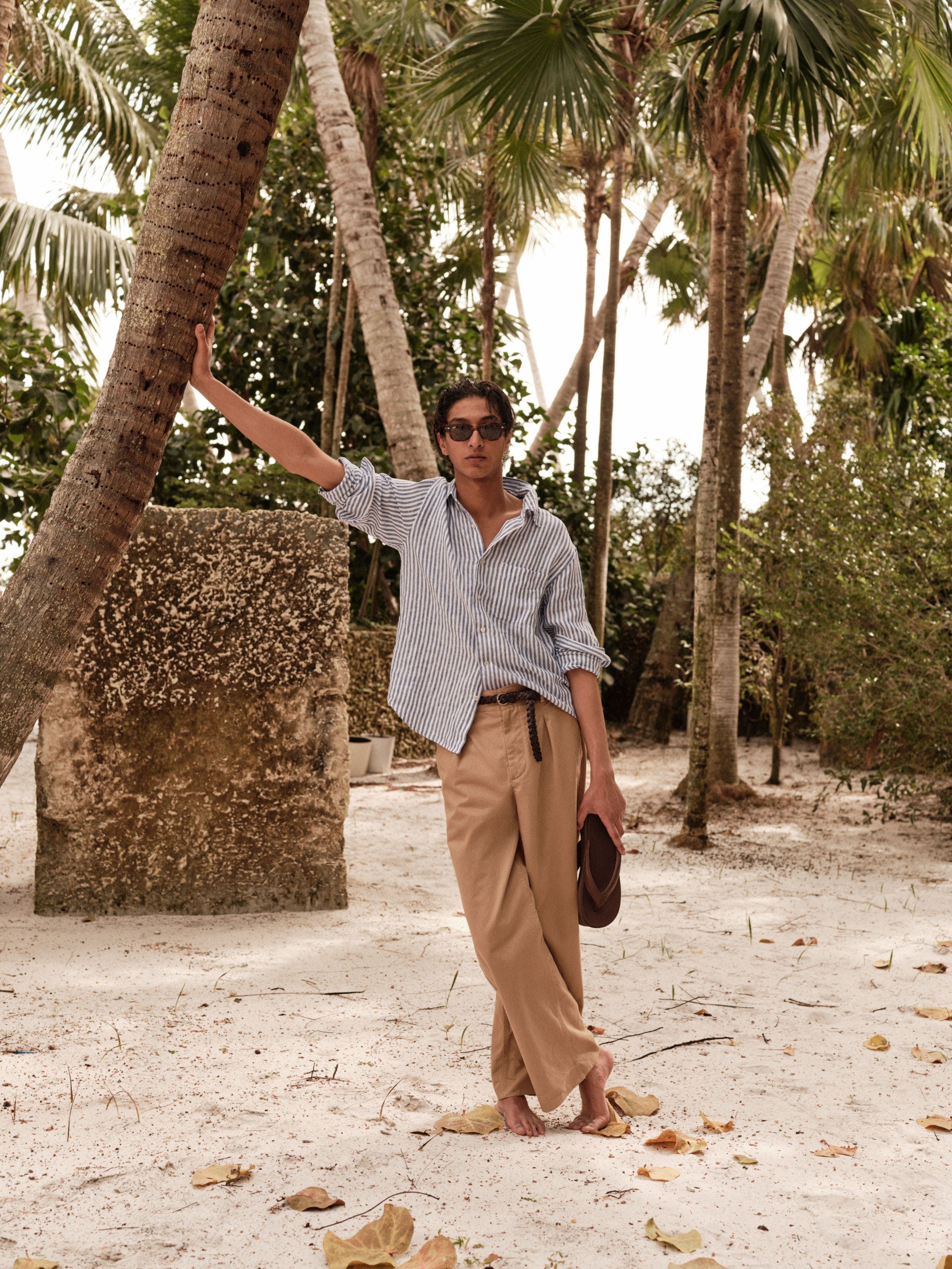 A man wearing sunglasses, a striped shirt, belt and khaki pants while holding flip-flops.