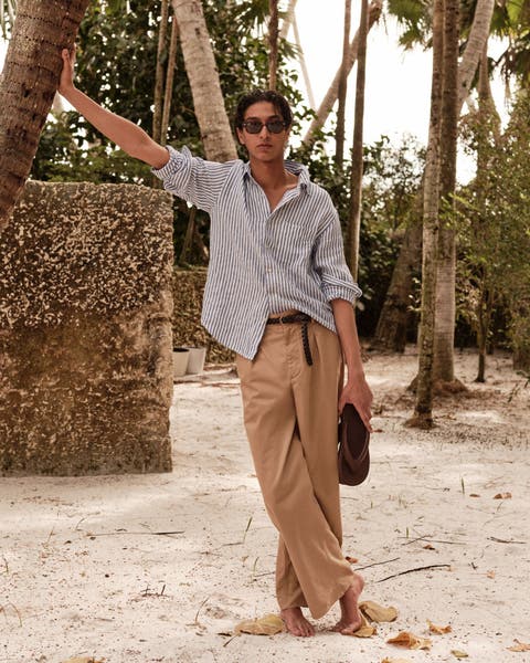 A man wearing sunglasses, a striped shirt, belt and khaki pants while holding flip-flops.