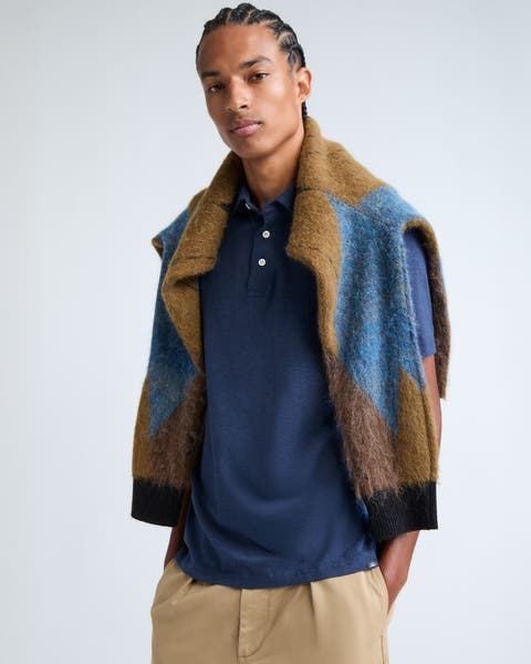 A man wearing a navy polo and khaki pants with a patterned sweater draped over his shoulders.