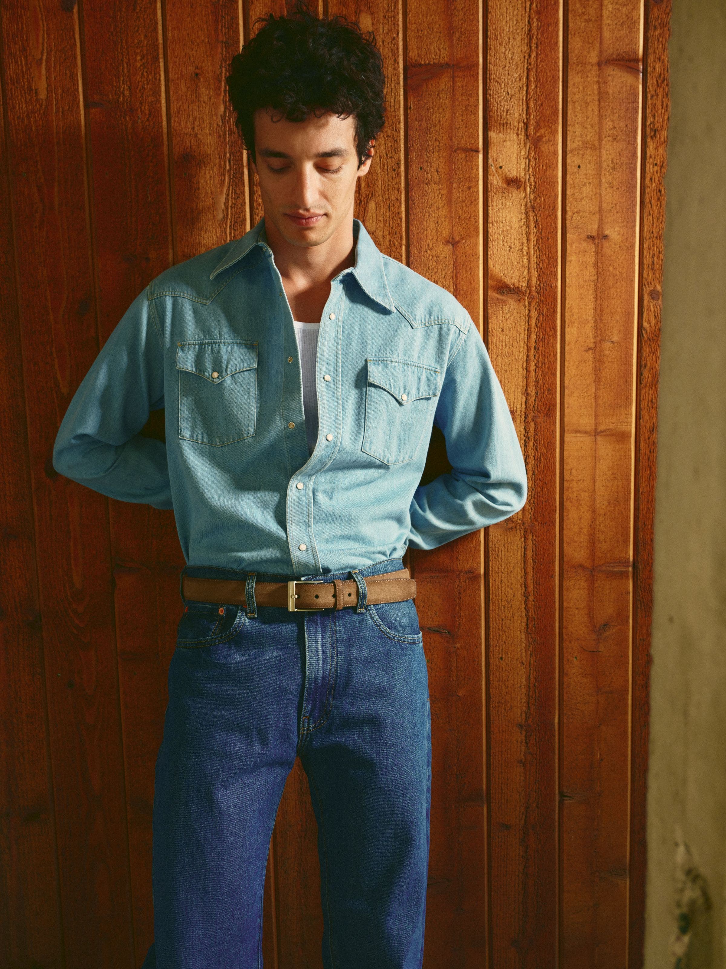 Man wearing a denim shirt over a tank with jeans and a belt.