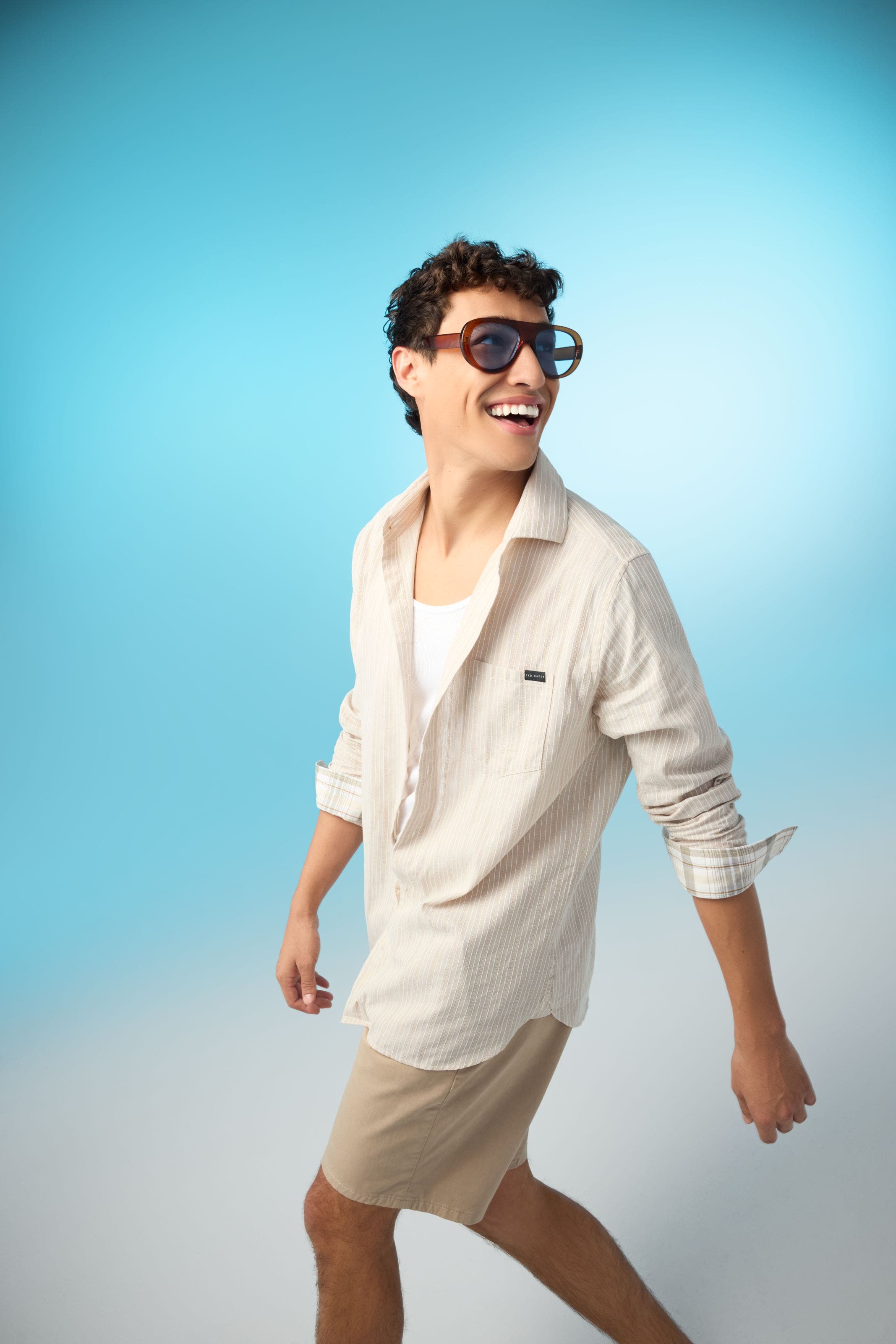 A man wearing a white tank, a striped cream shirt and tan shorts.