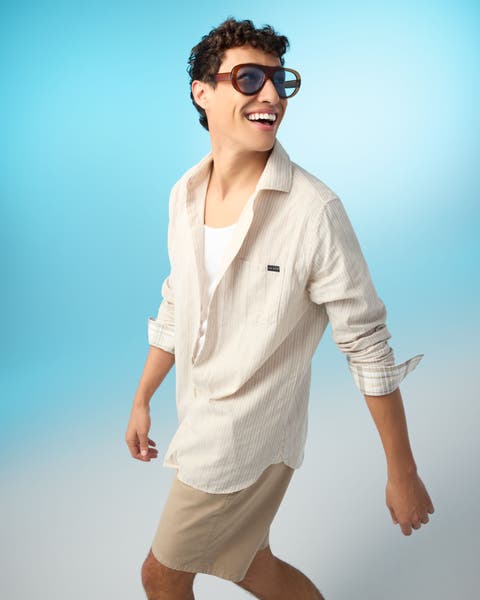 A man wearing a white tank, a striped cream shirt and tan shorts.