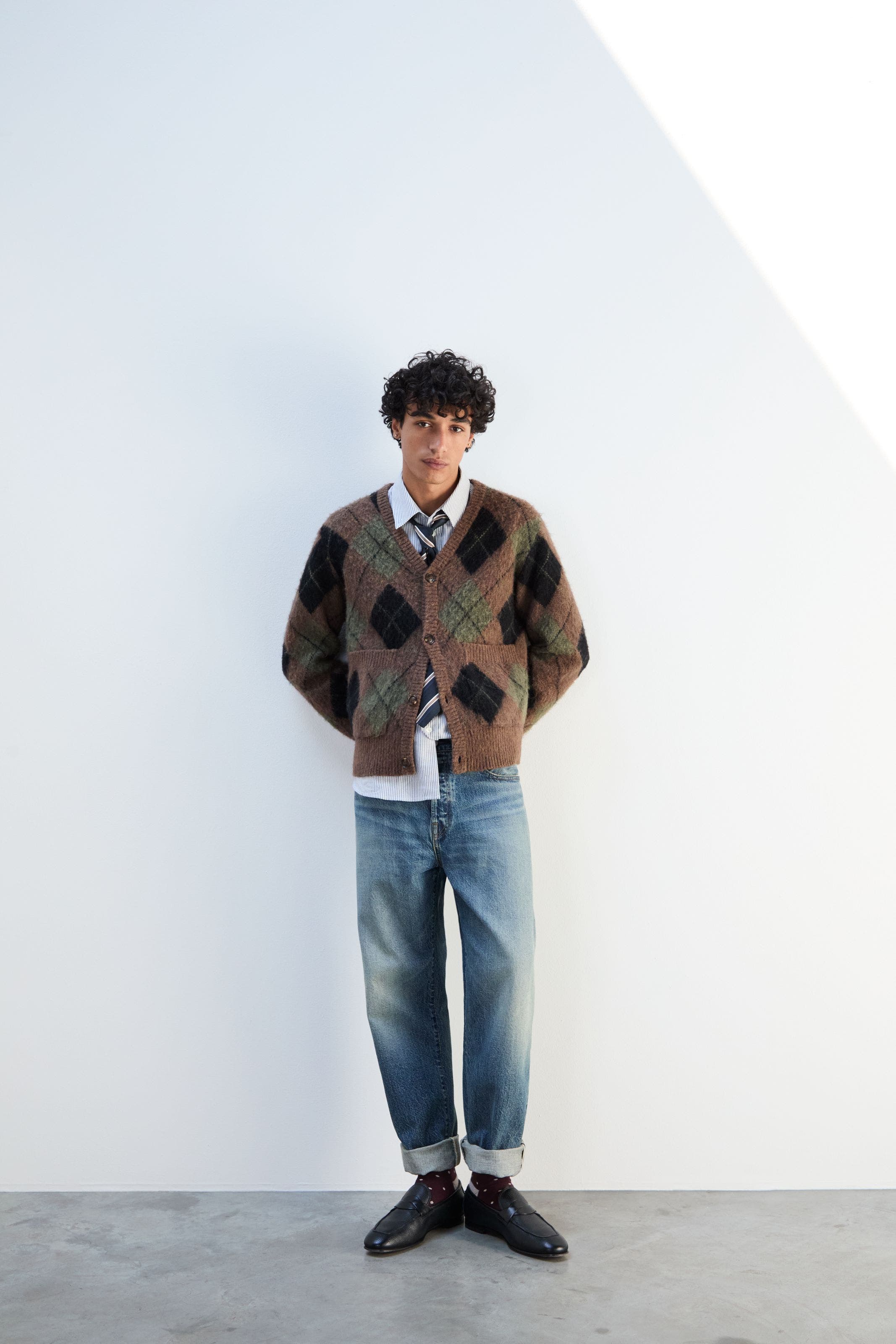 A man wearing an argyle cardigan and jeans.