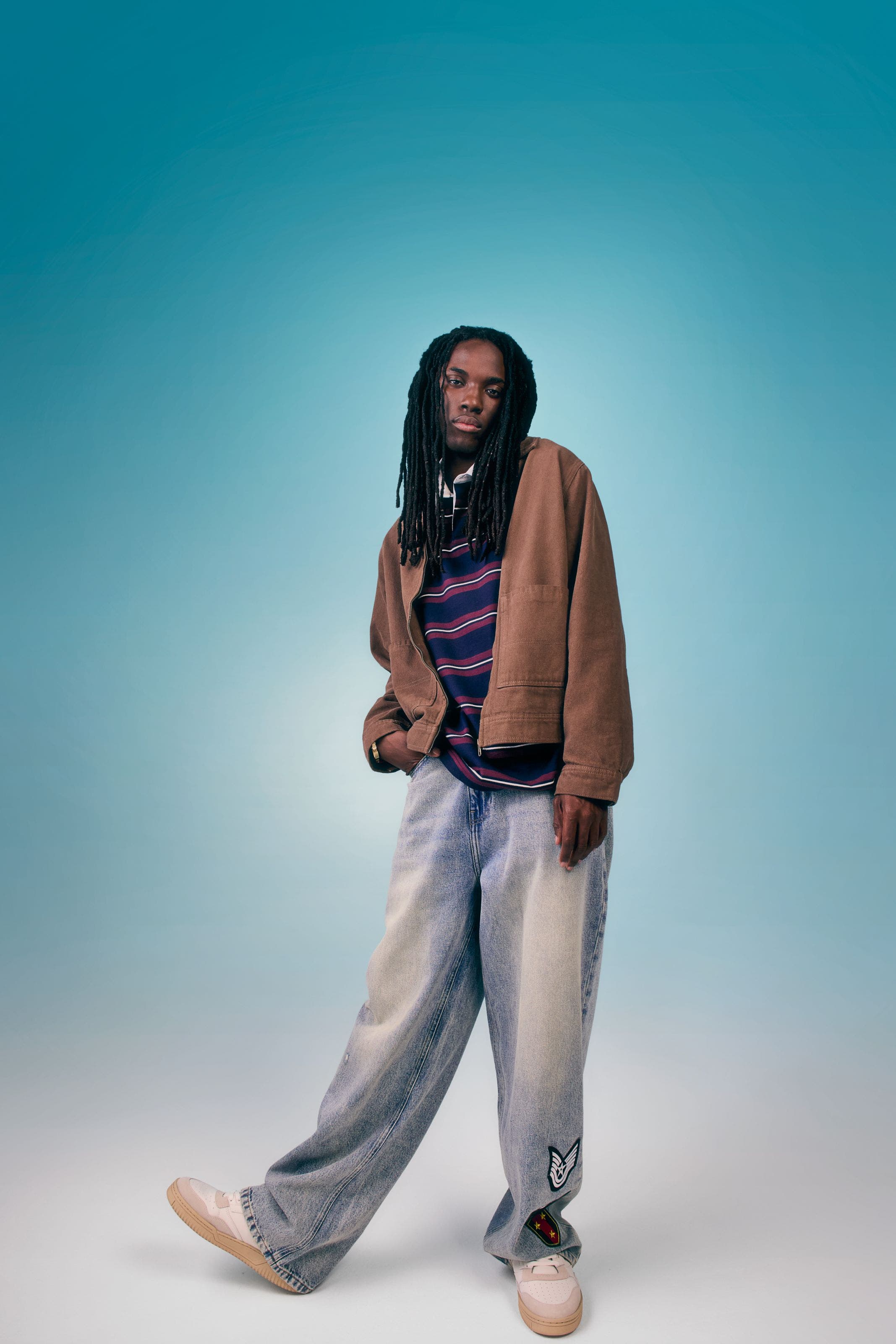 Male model wearing a tan bomber jacket over a navy striped sweater with light wash jeans.