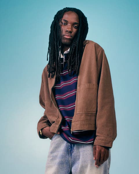 Male model wearing a tan bomber jacket over a navy striped sweater with light wash jeans.