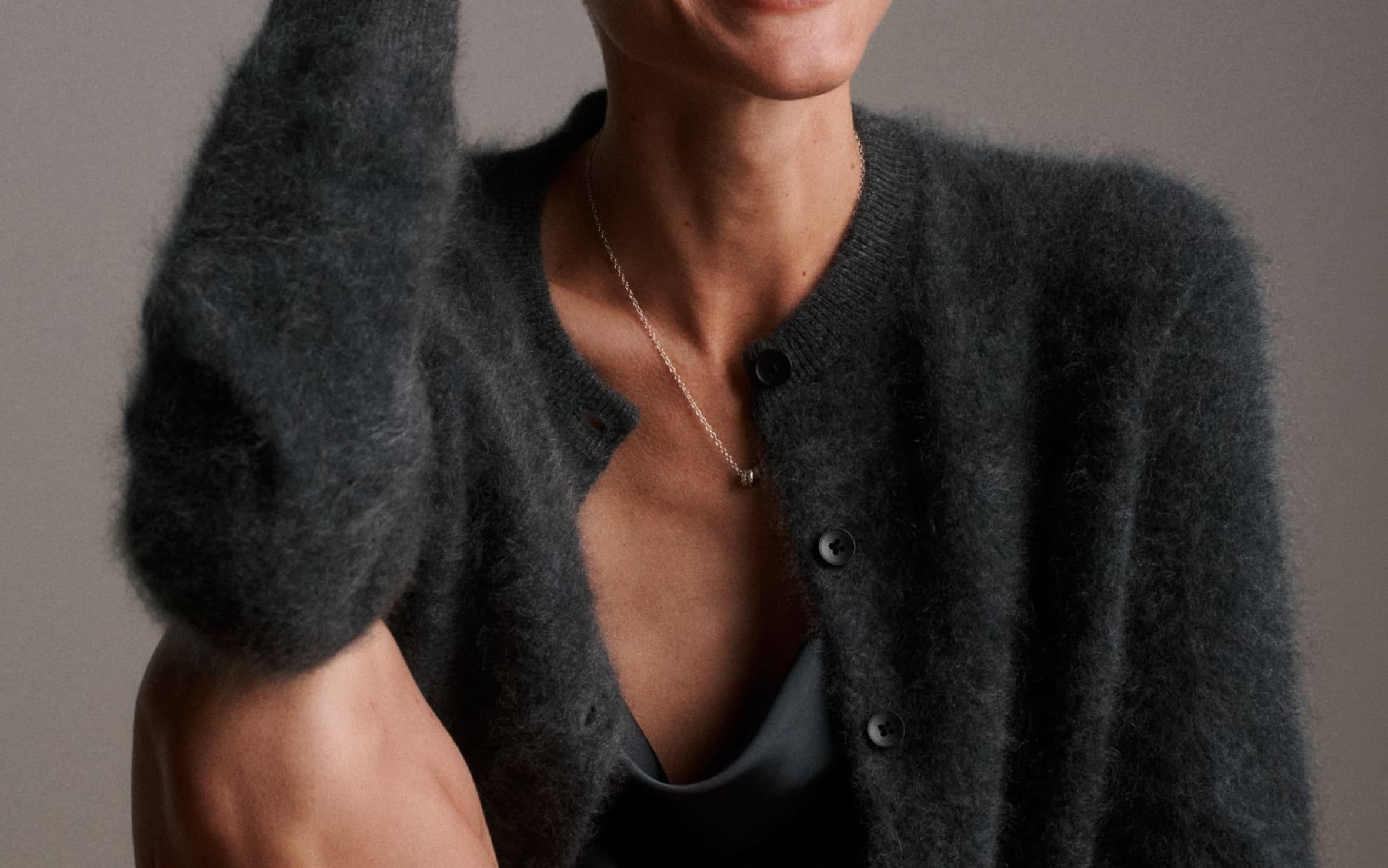 A woman wearing a Spinelli Kilcollin pendant necklace, earrings and stacked rings with a fuzzy black cardigan and satin cowl-neck top.