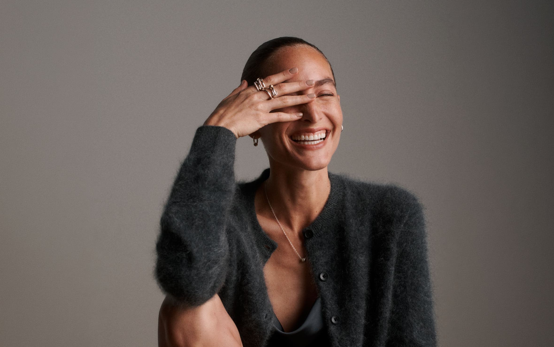 A woman wearing a Spinelli Kilcollin pendant necklace, earrings and stacked rings with a fuzzy black cardigan and satin cowl-neck top.