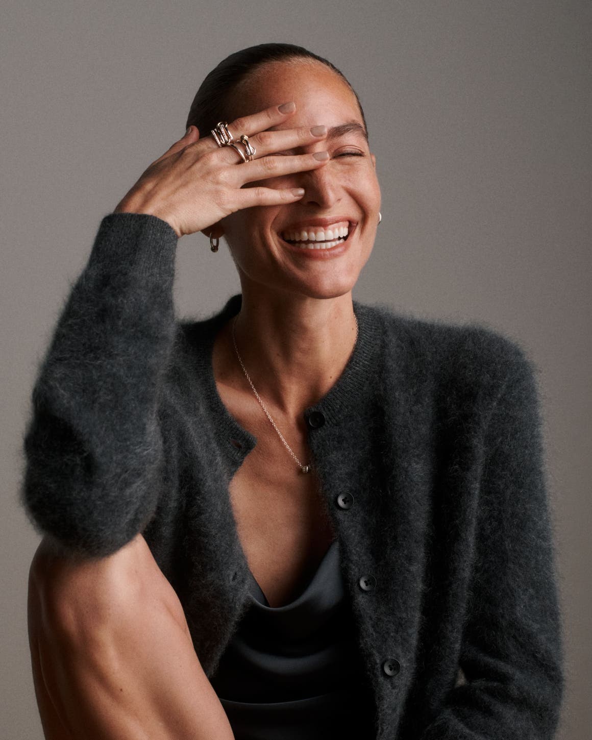 A woman wearing a Spinelli Kilcollin pendant necklace, earrings and stacked rings with a fuzzy black cardigan and satin cowl-neck top.