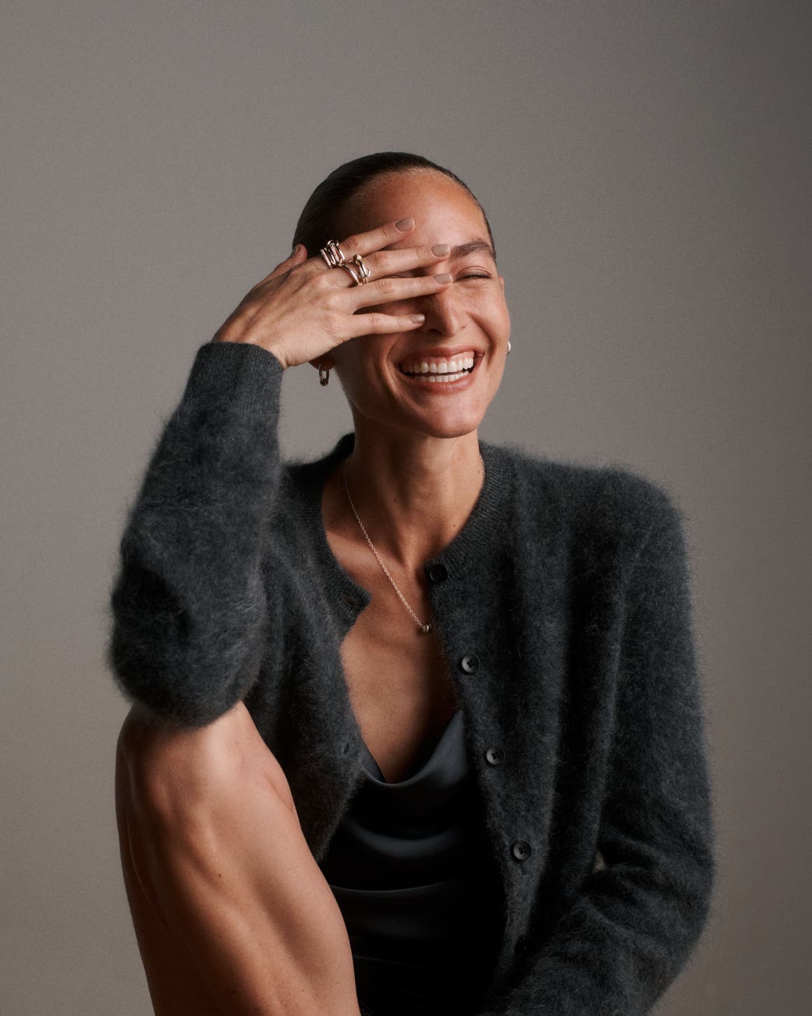 A woman wearing a Spinelli Kilcollin pendant necklace, earrings and stacked rings with a fuzzy black cardigan and satin cowl-neck top.