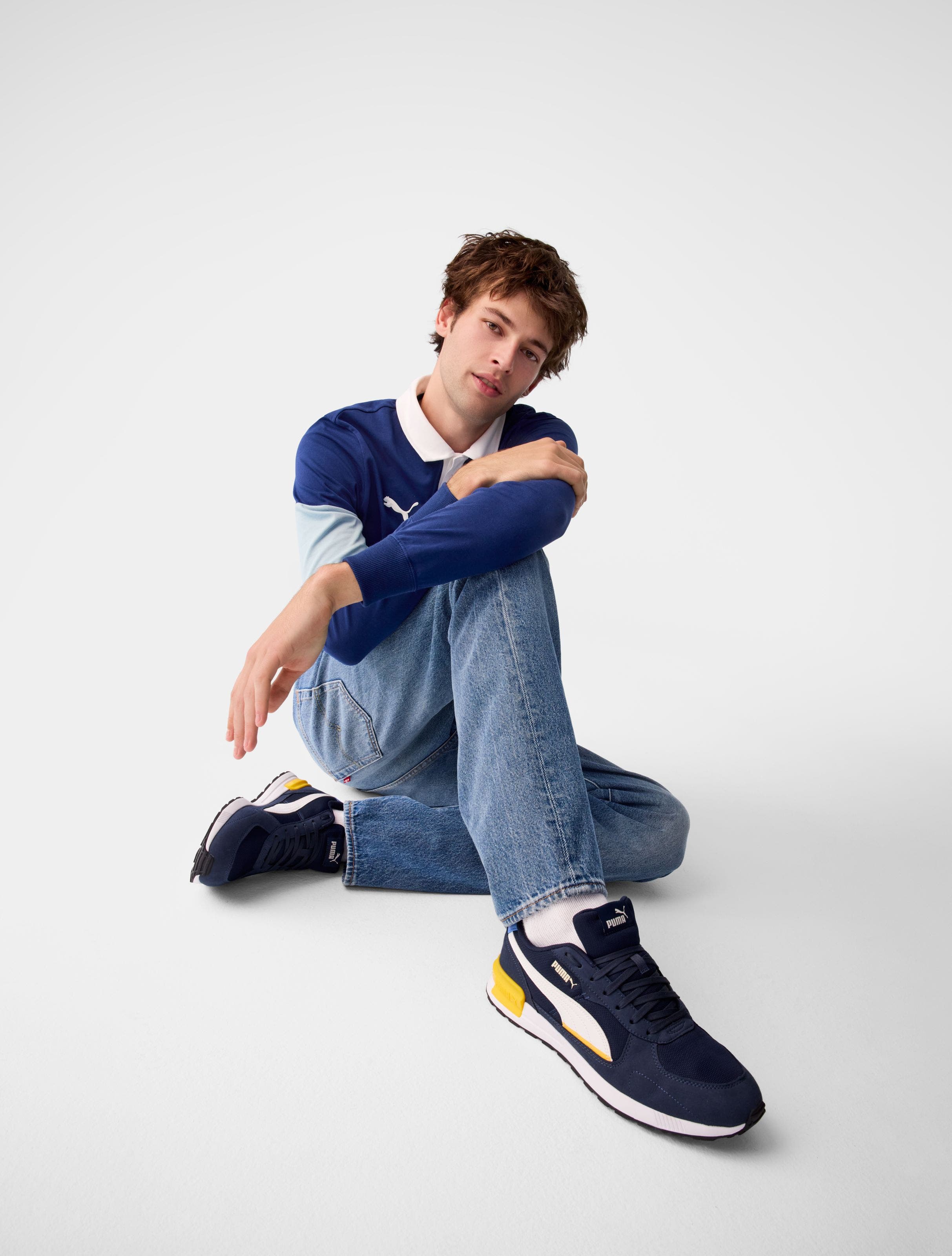 A man wearing a blue long-sleeve polo with jeans and navy sneakers.