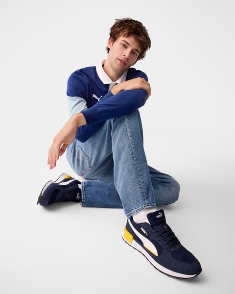 A man wearing a blue long-sleeve polo with jeans and navy sneakers.