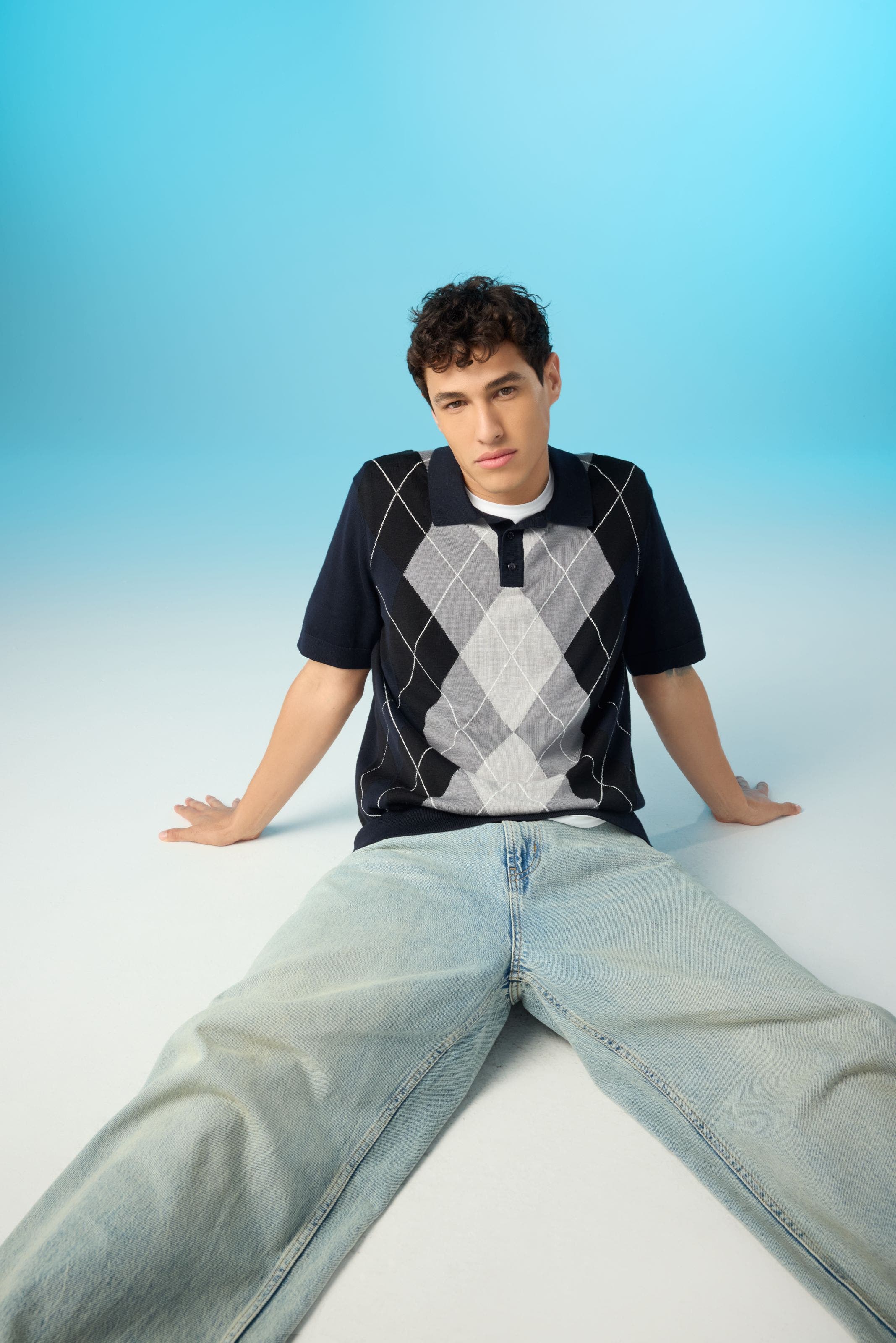 A man wearing a knit argyle top, a white tee and light-wash jeans. 