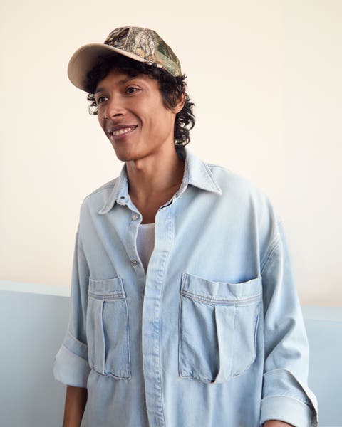 A man wearing a denim button up long sleeve shirt and cap.