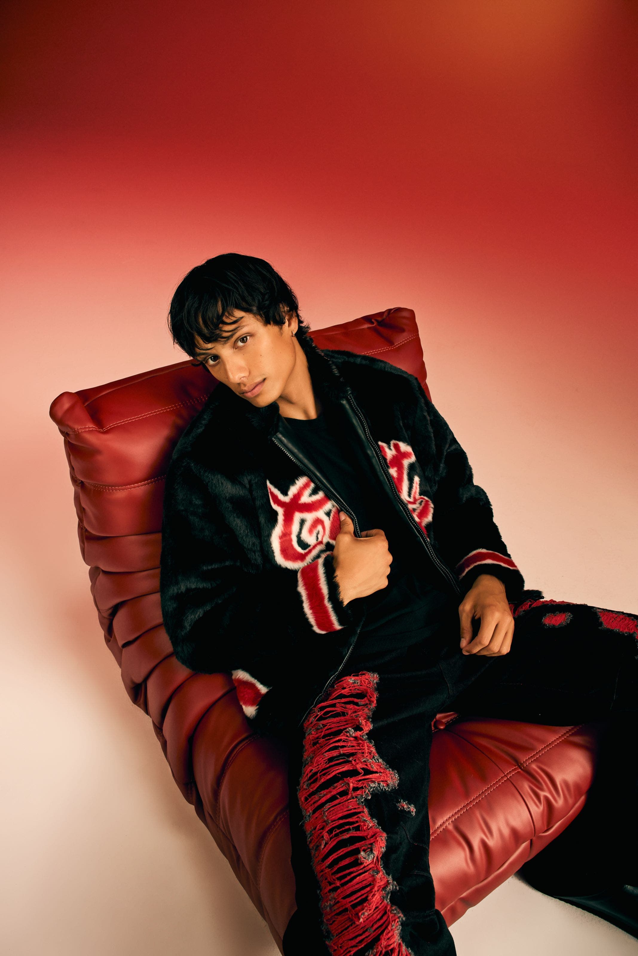 Man in a graphic jacket and distressed pants on a red leather chair.