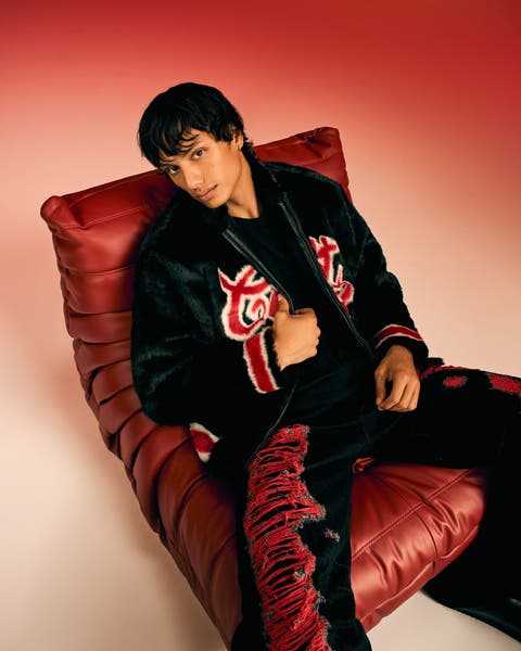 Man in a graphic jacket and distressed pants on a red leather chair.