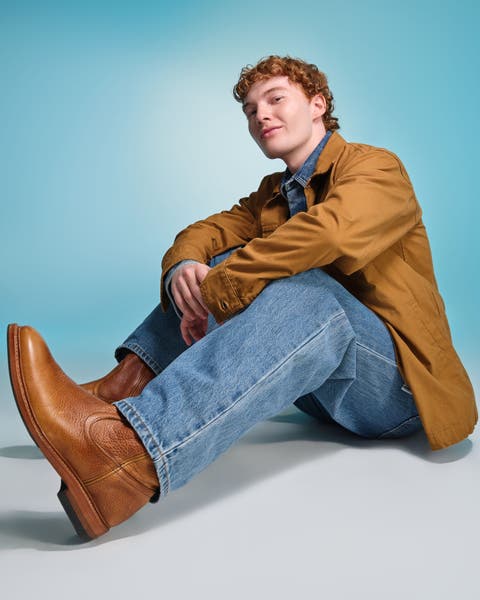 Man wearing a tan jacket, blue jeans, and brown leather boots.