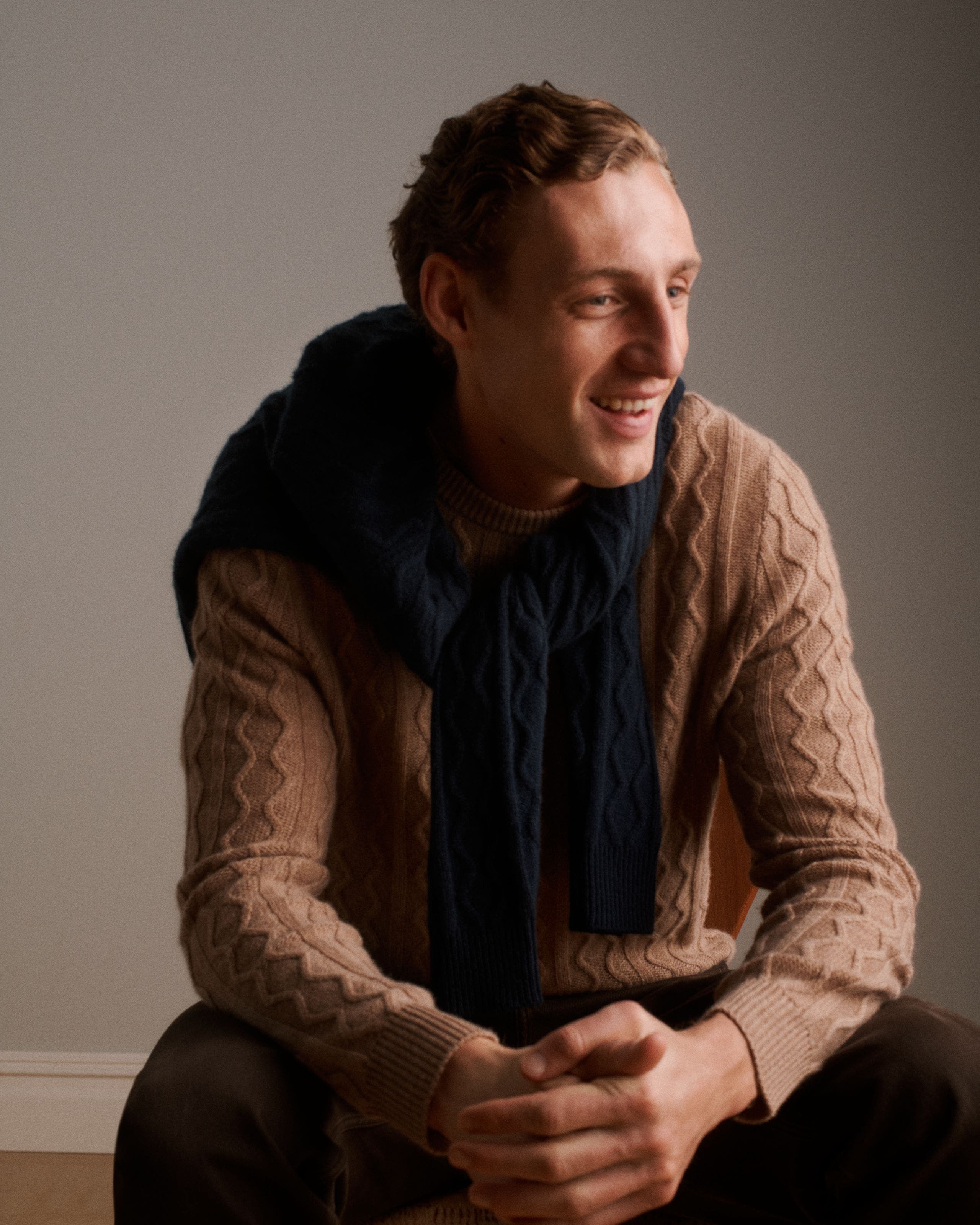 A man wearing a textured camel crewneck sweater, a navy knit layered over his shoulders.