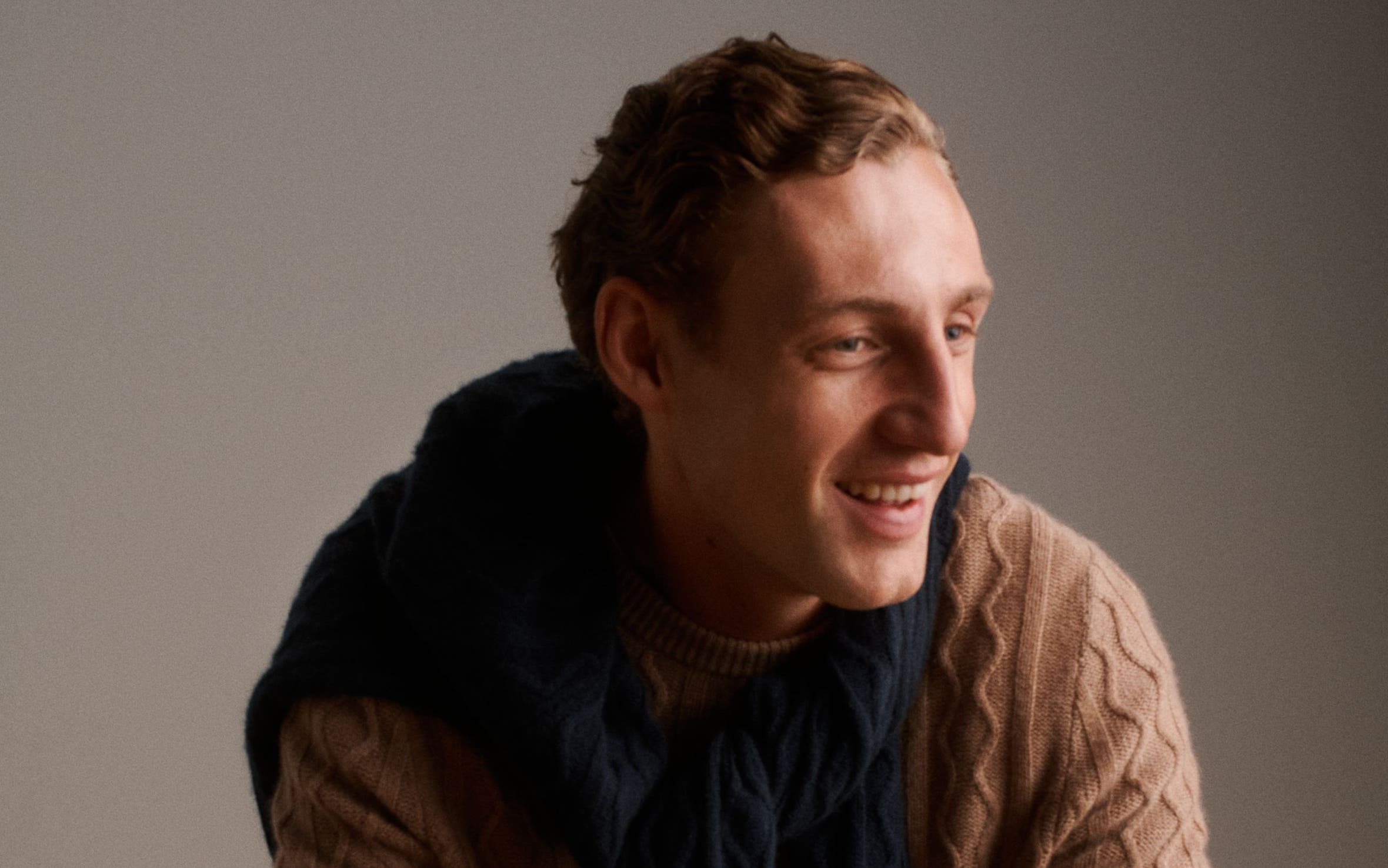A man wearing a textured camel crewneck sweater, a navy knit layered over his shoulders.