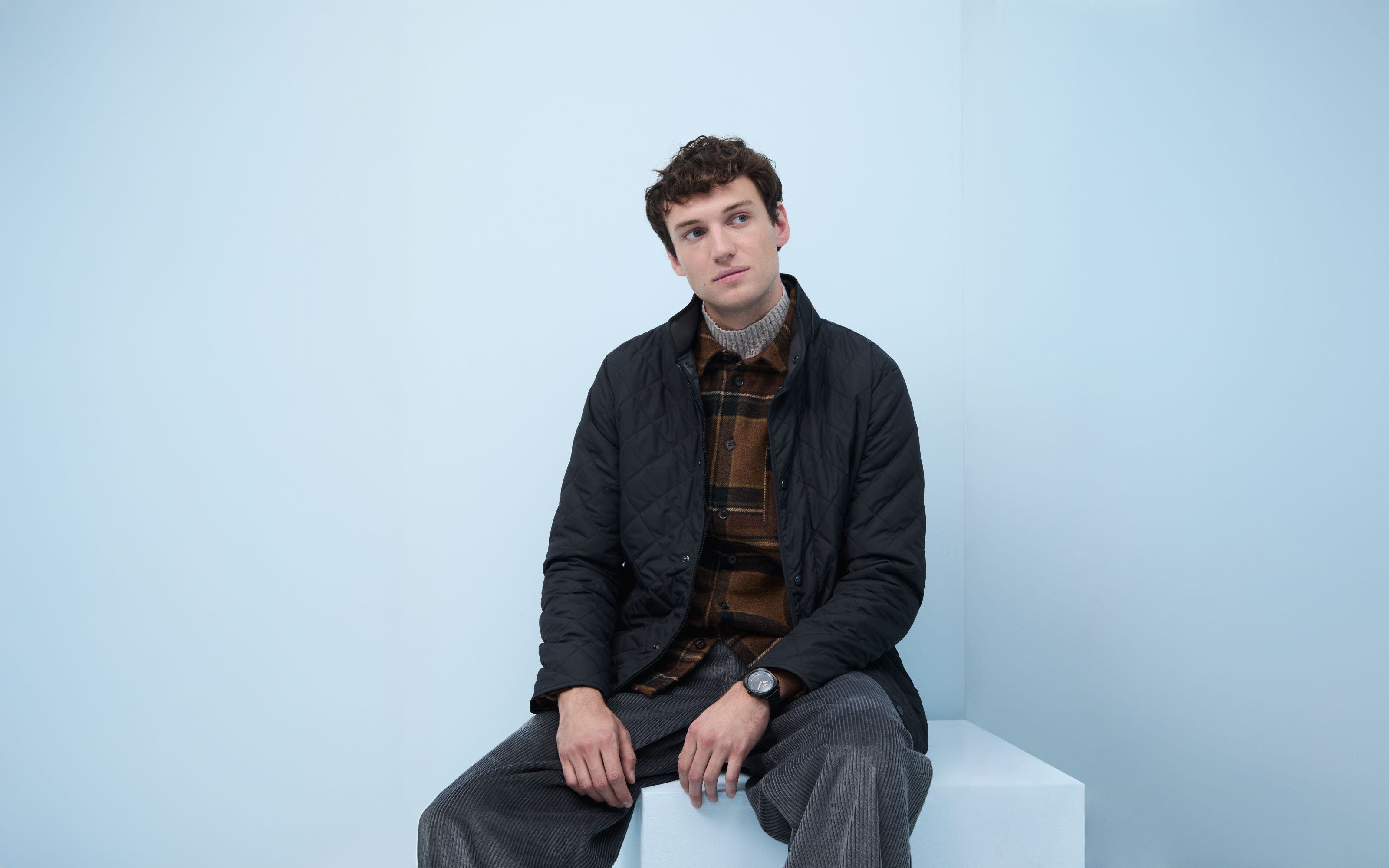 A man wearing a quilted jacket, plaid overshirt, turtleneck sweater and corduroy pats.