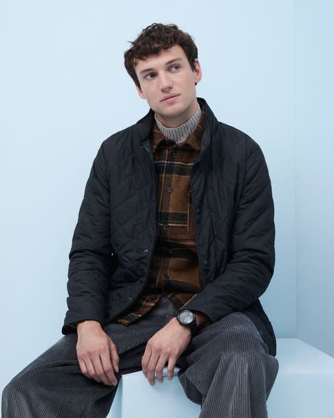 A man wearing a quilted jacket, plaid overshirt, turtleneck sweater and corduroy pats.