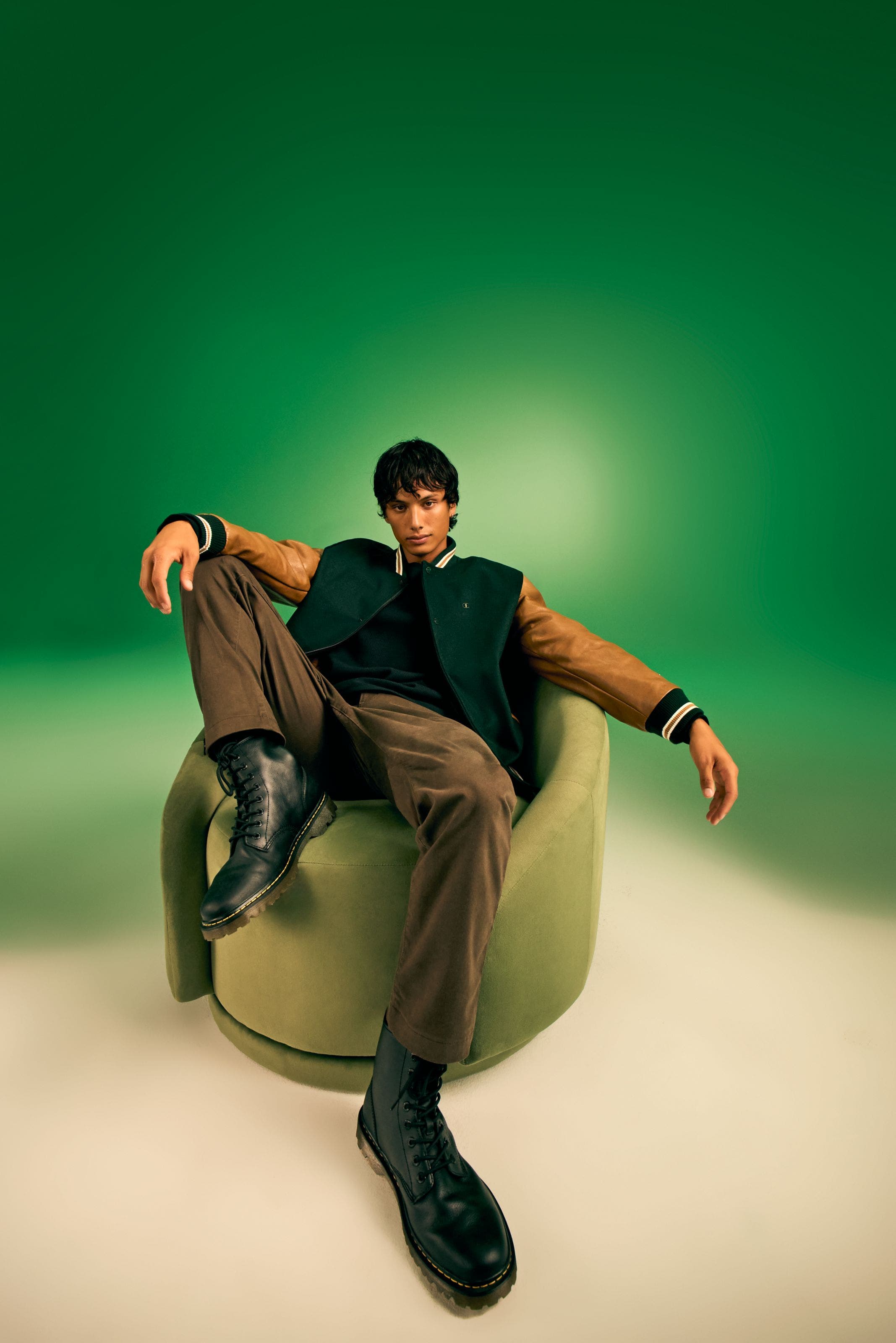 Male model wearing a varsity jacket, brown trousers, and black lace-up boots.