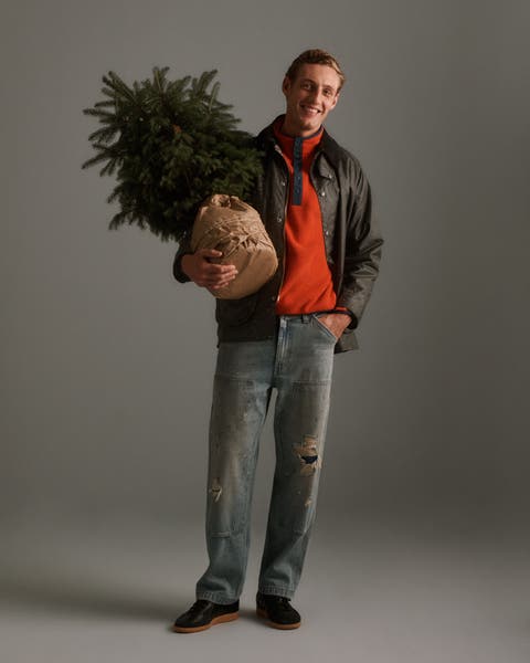 A man wearing a jacket, sweater, and jeans holding a tree.