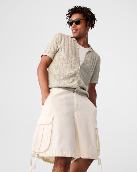 A man wearing a white tank, a tan cardigan and cream cargo shorts.