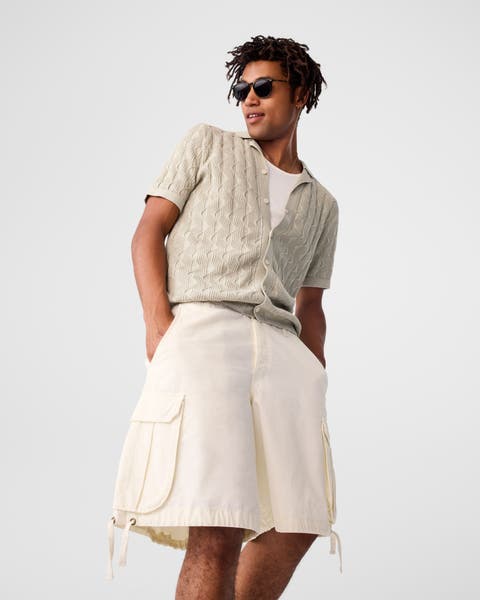 A man wearing a tan cardigan and cream cargo shorts.