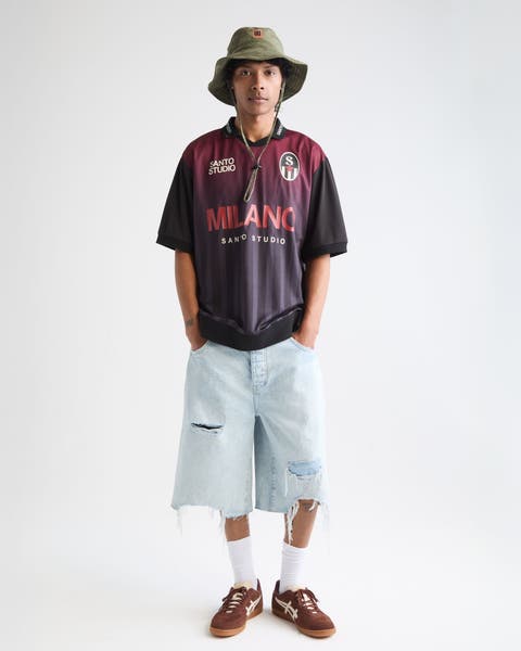 Man wearing a soccer jersey, jean shorts, bucket hat and sneakers.