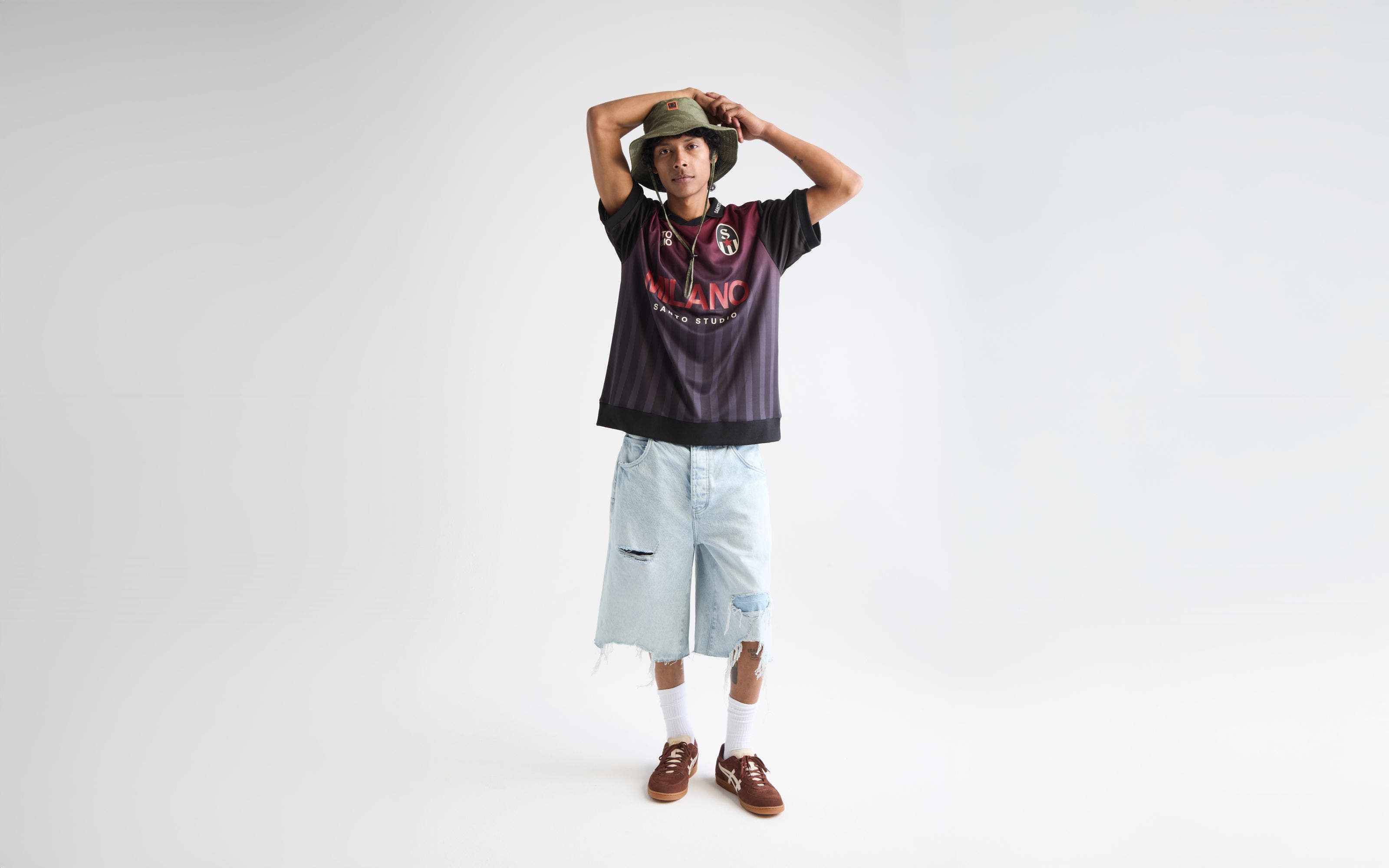 Man wearing a soccer jersey, jean shorts, bucket hat and sneakers.