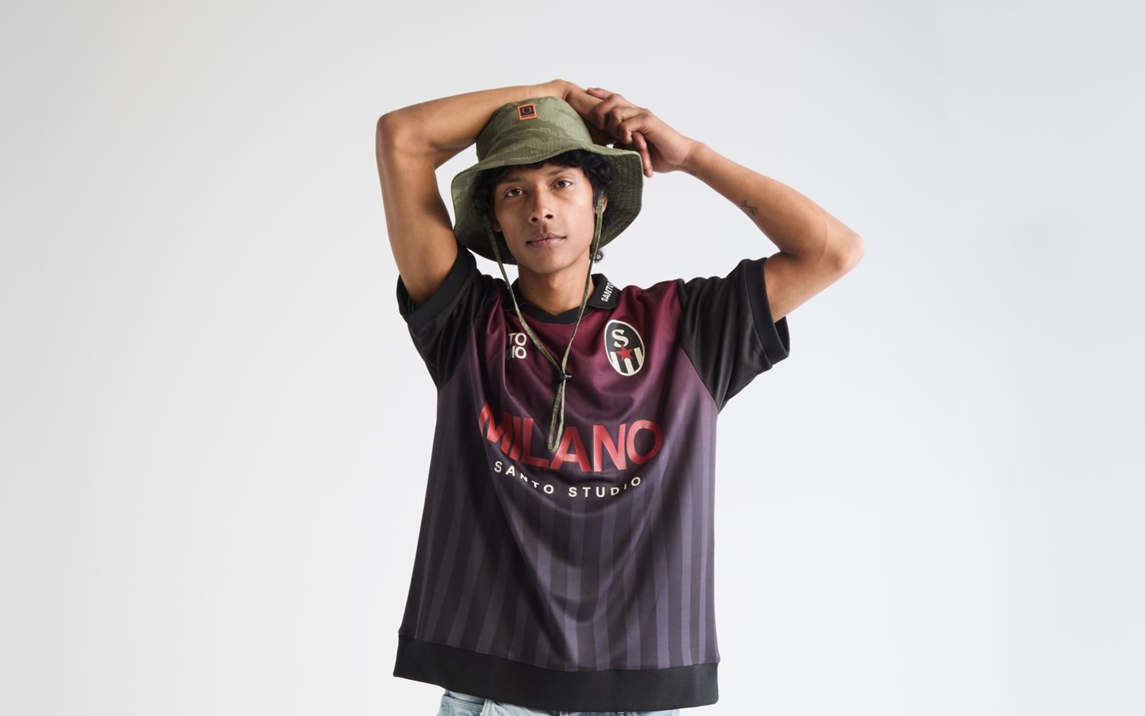 Man wearing a soccer jersey, jean shorts, bucket hat and sneakers.