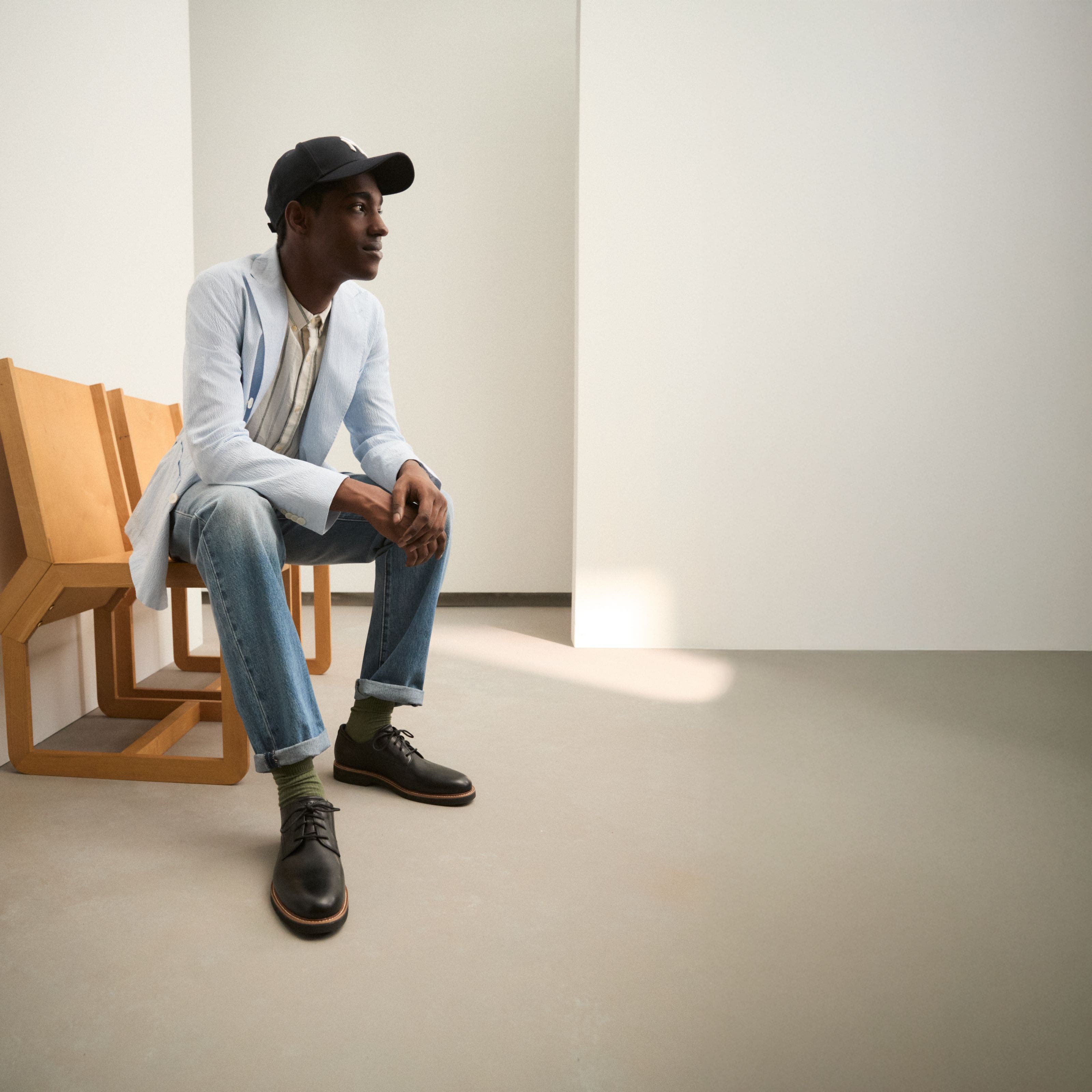 A man wearing a lightweight sport coat, button-down shirt, jeans and ball cap.